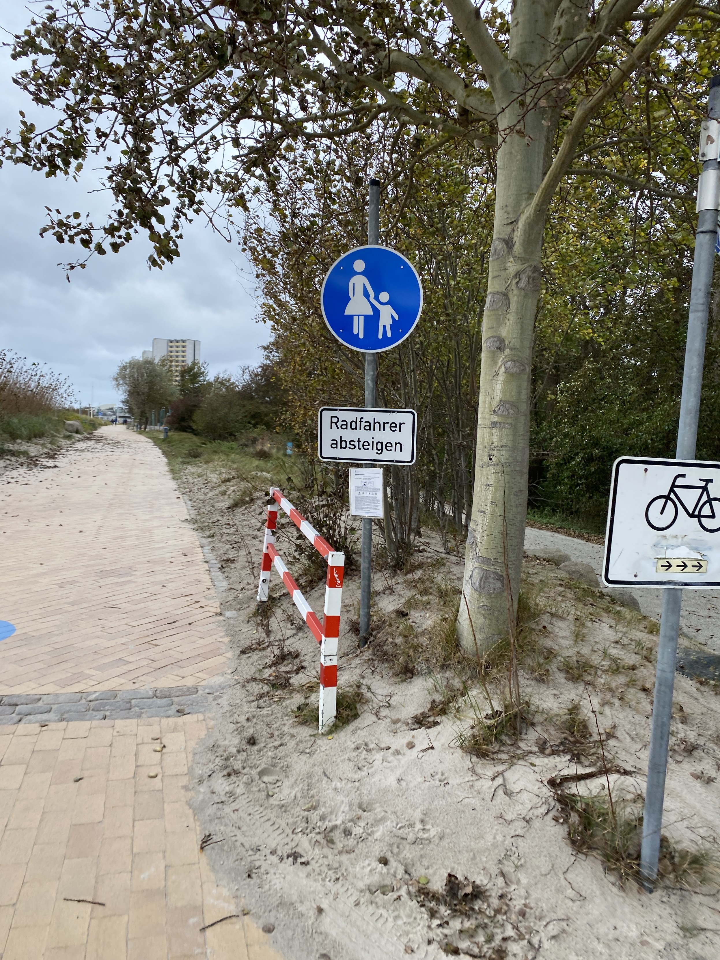 Eine Szene an der Ostsee; Strandpromenade. Geradeaus ein Schild Gehweg mit Zusatzschild „Radfahrer Absteigen“. Am rechten Bildrand ein Schild mit Fahrradpiktogramm und kleinen Pfeilen nach rechts. Geradeaus auf dem Gehweg weit und breit kaum Personen zu sehen.
