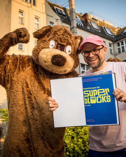 Foto von Bärenkostüm und Person, die die Druckversion der Empfehlungen für Superblocks in der Hand hält und lacht.