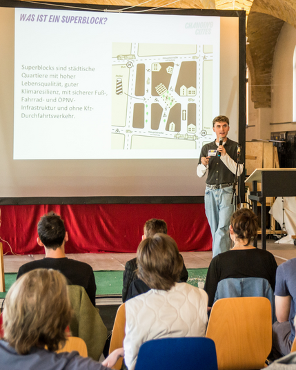 Foto der Veranstaltung, auf der Bühne steht eine Person mit Mikrofon. Auf der Präsentation dahinter steht geschrieben: Was ist ein Superblock? Superblocks sind städtische Quartiere mit hoher Lebensqualität, guter Klimaresilienz, mit sicherer Fuß-, Fahrrad- und ÖPNV-Infrastruktur und ohne KFZ-Durchfahrtsverkehr.